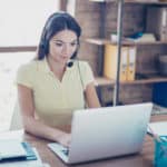 Successful young Latin girl is talking to customer with headset on and putting data in her computer